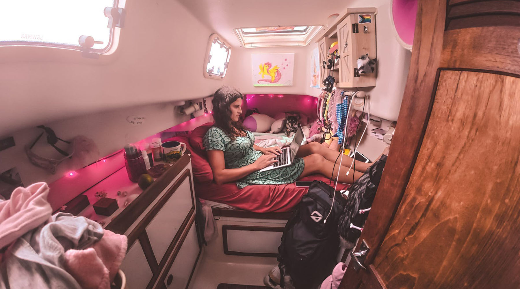 Kelsey Granger sits on a bed in a small, cozy boat cabin with pink lights and decorations, typing on a laptop with a dog lying next to her.