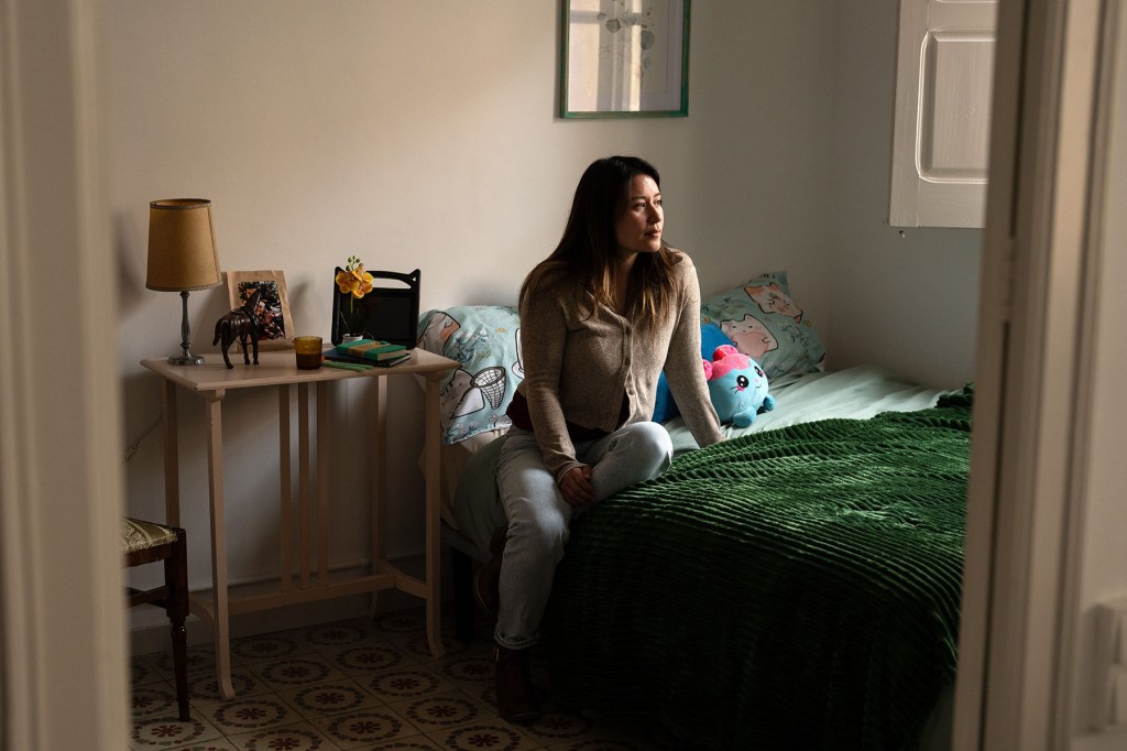 Jewel Lazaro sits on a bed in a room decorated with stuffed animals and colorful bedding, looking out toward the window.