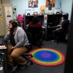 A person sits at a desk on the phone in an office with others on phones and a rainbow rug on the floor.