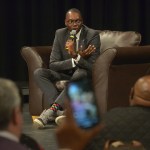 Garlin Gilchrist sits onstage in a suit and sneakers, speaking to a crowd.